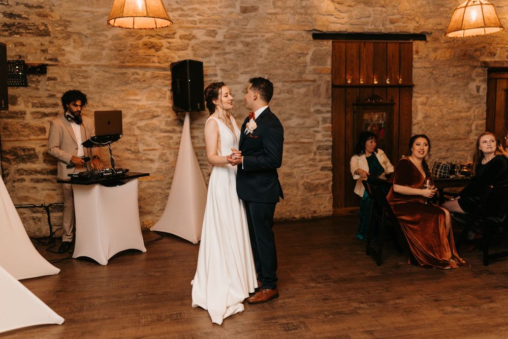 Bride and groom share their first dance in a warmly lit rustic stone ballroom with guests watching.