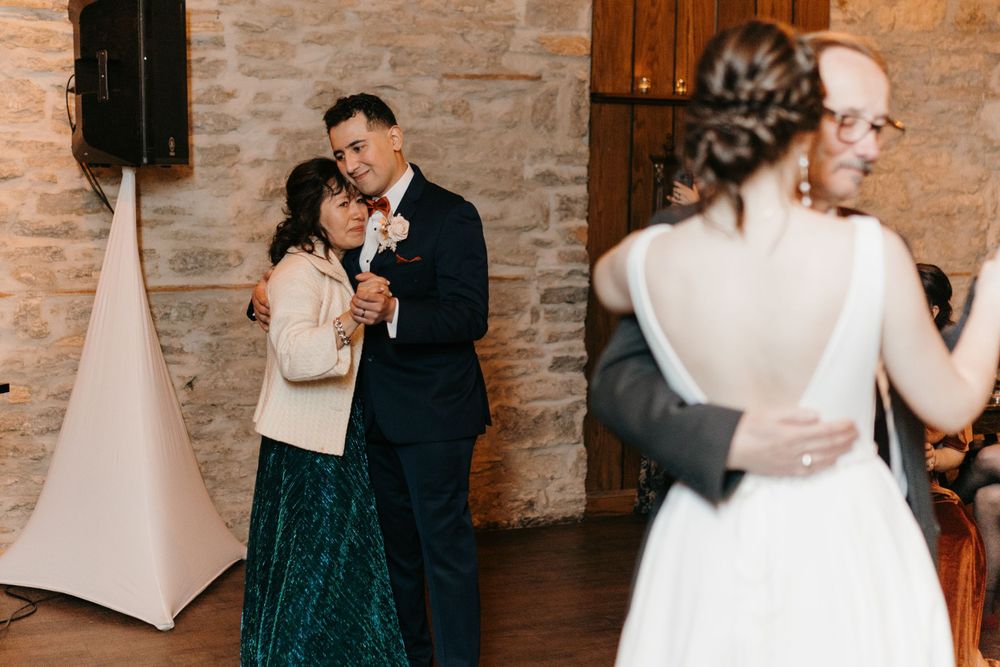 Groom dances with his mother in a teal dress while bride dances with her father in the foreground.