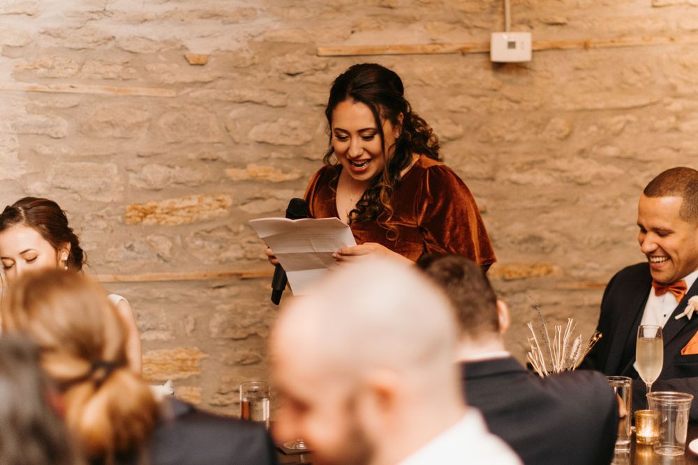 Bridesmaid in velvet dress delivering a heartfelt speech at a rustic wedding reception.