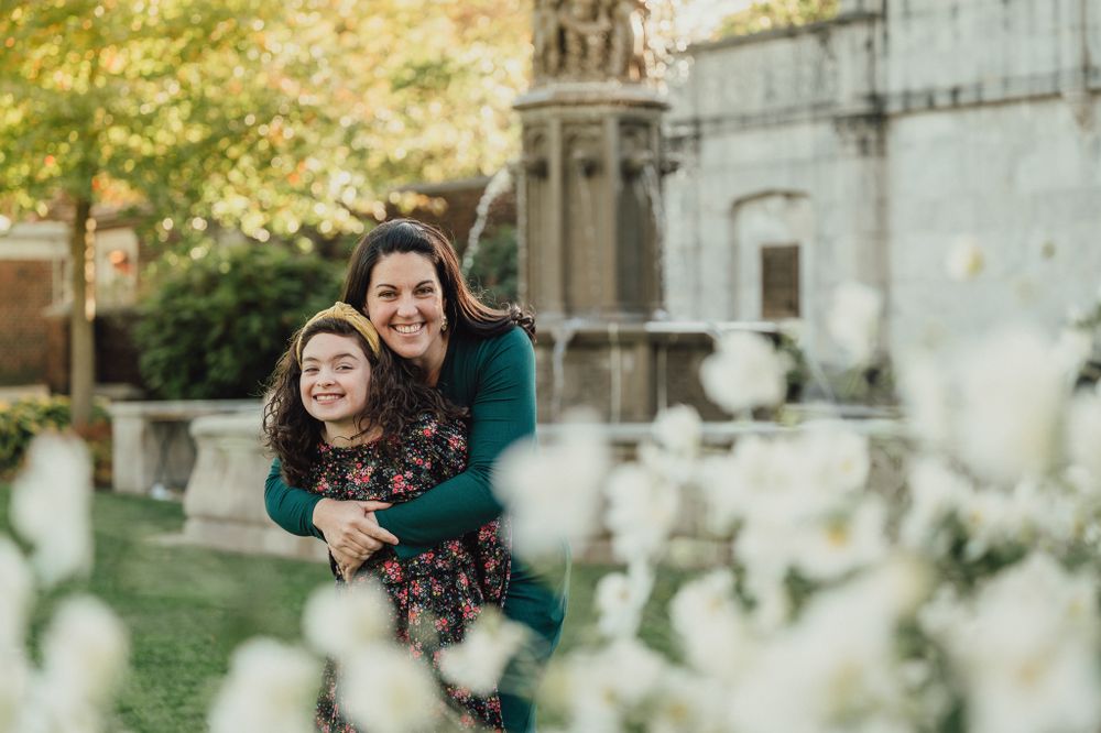 A smiling mother and daughter embrace warmly in front of white flowers and a stone fountain outdoors.