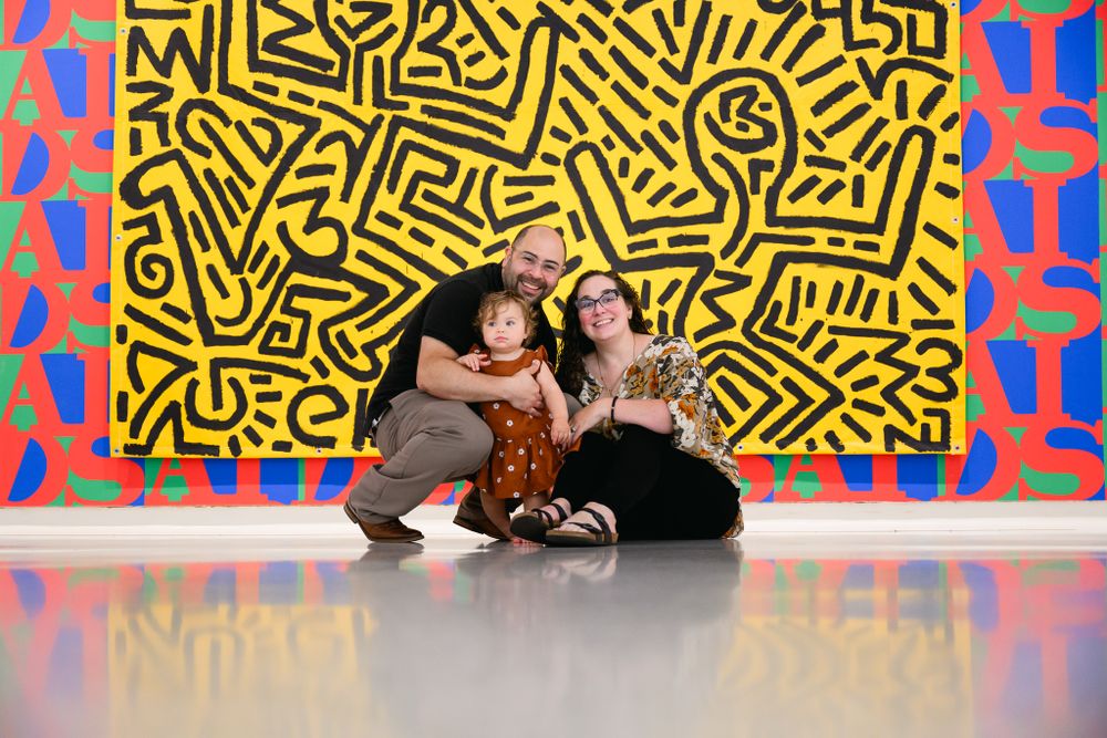 Happy family of three sits on museum floor before colorful Keith Haring artwork.