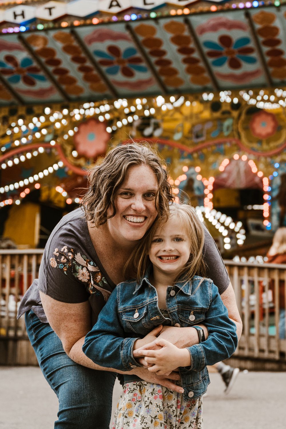 Mother hugging daughter in front of colorful lit carousel at amusement park, both smiling.