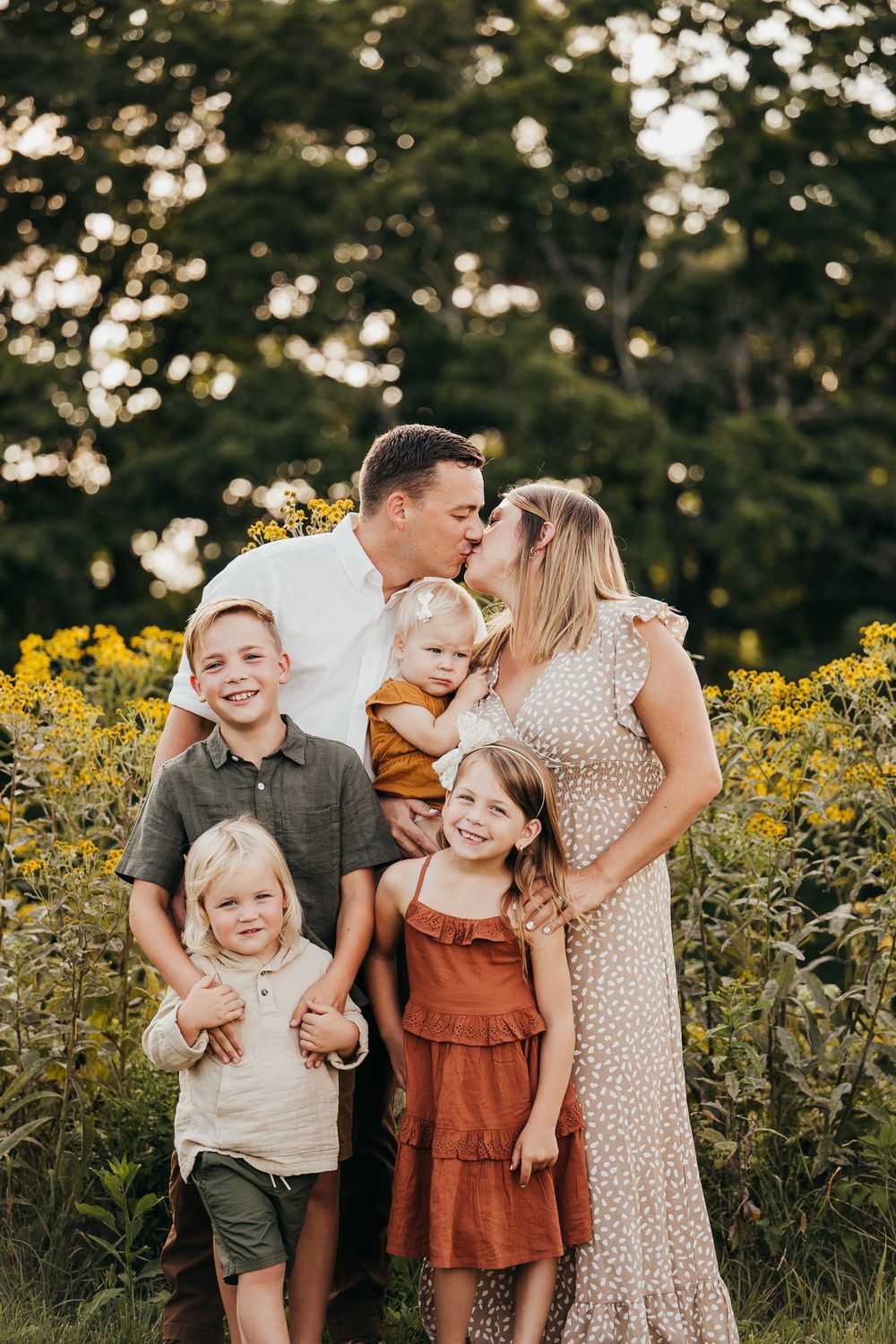 Family of six poses in sunlit wildflower field, parents kissing with four children.