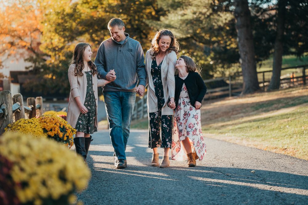 A joyful family of four walks together along a sunlit autumn path lined with yellow chrysanthemums.