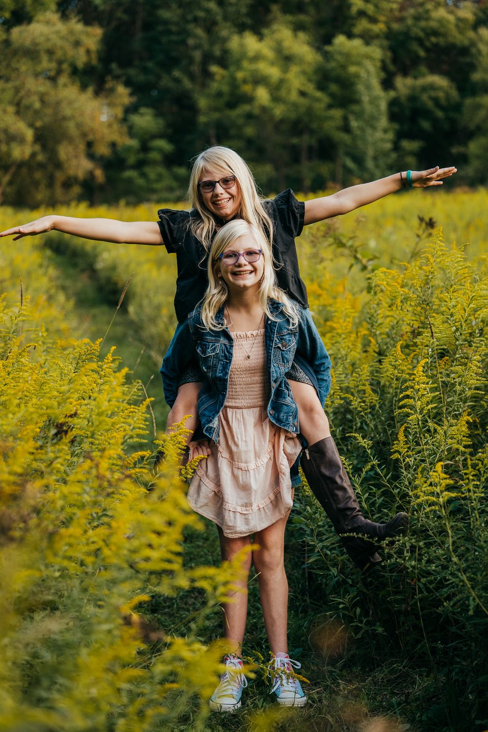 A blonde mother sits on her daughter's shoulders with arms outstretched in a vibrant yellow wildflower field.