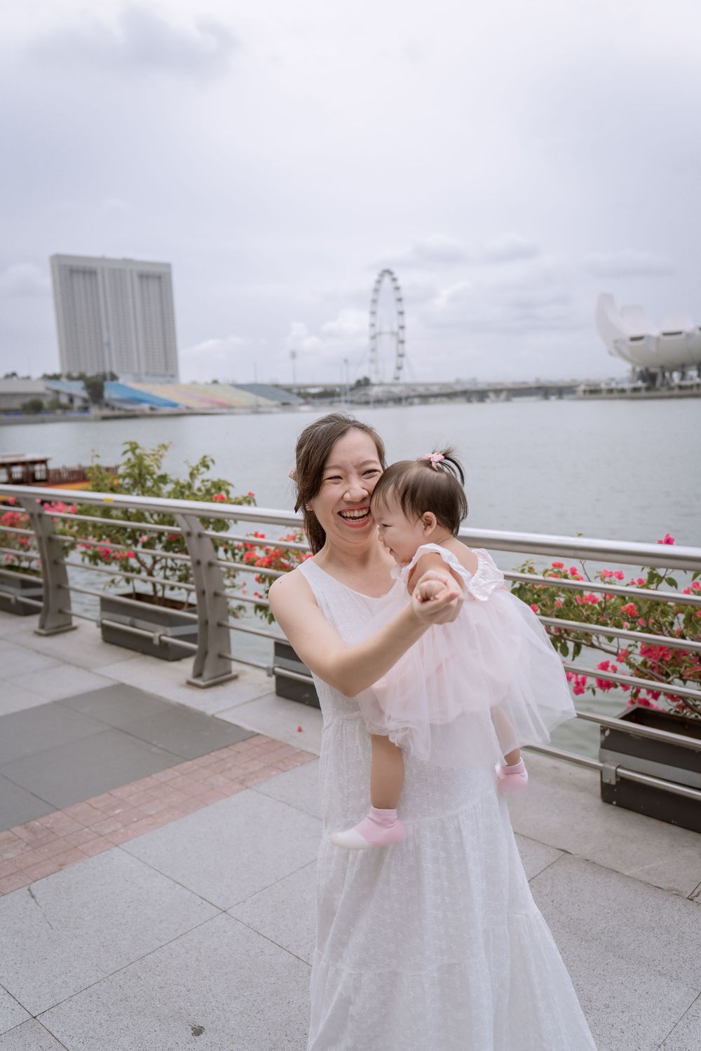 Mother and daughter in white dresses share a joyful moment by the waterfront with Singapore Flyer in background.