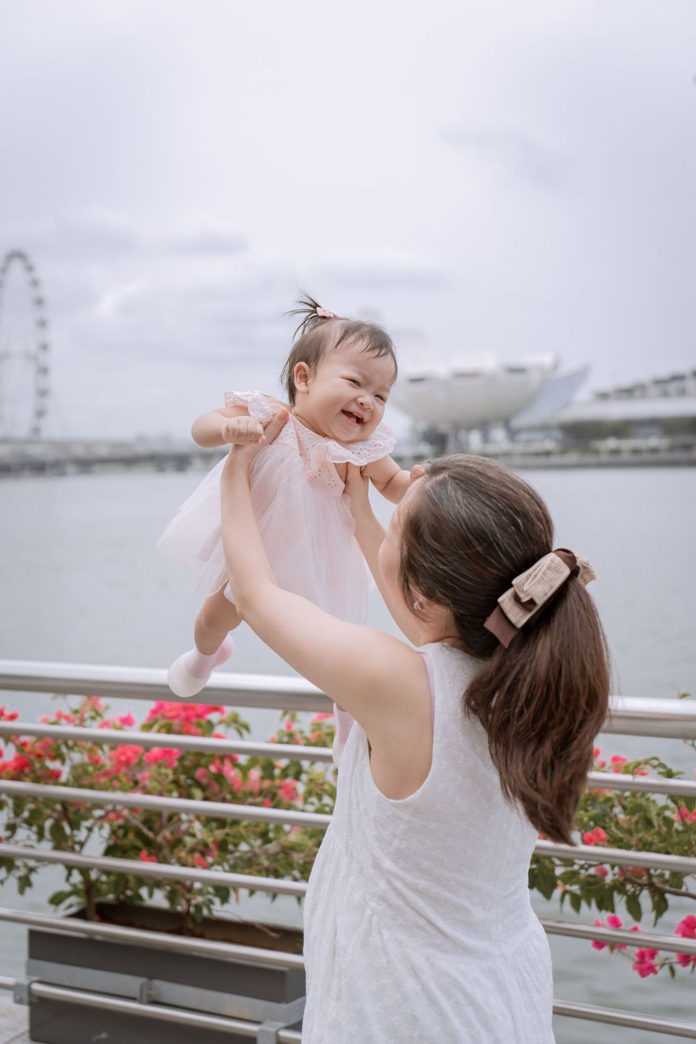 Baby in pink dress being lifted up with Singapore skyline and Ferris wheel in background.