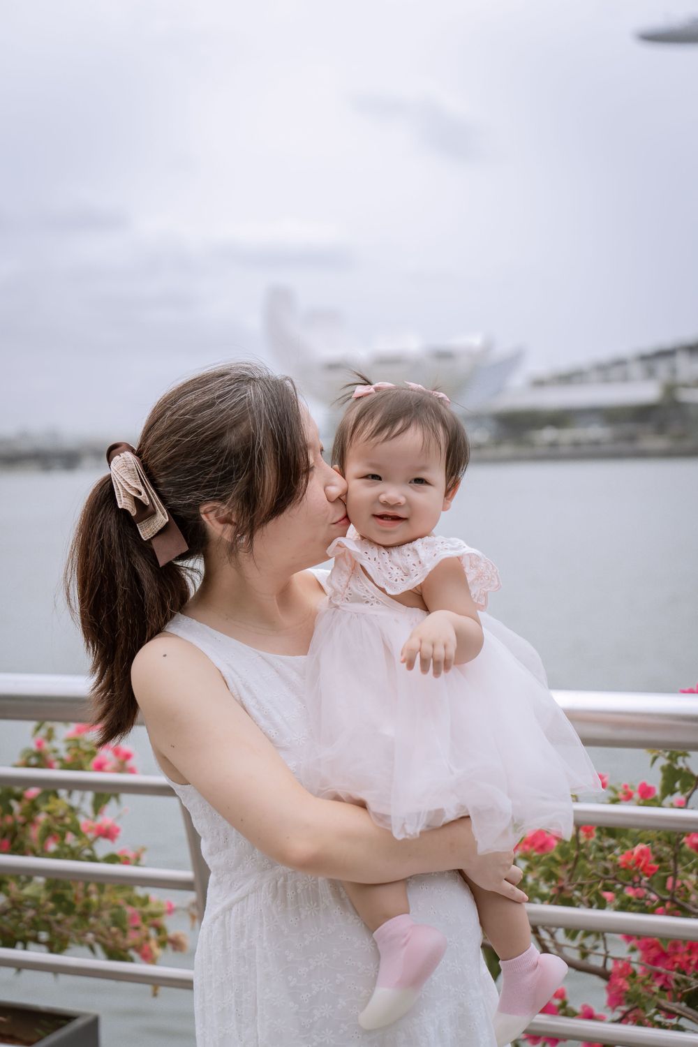 Parent holding smiling baby in white dress with Marina Bay Sands visible behind them.