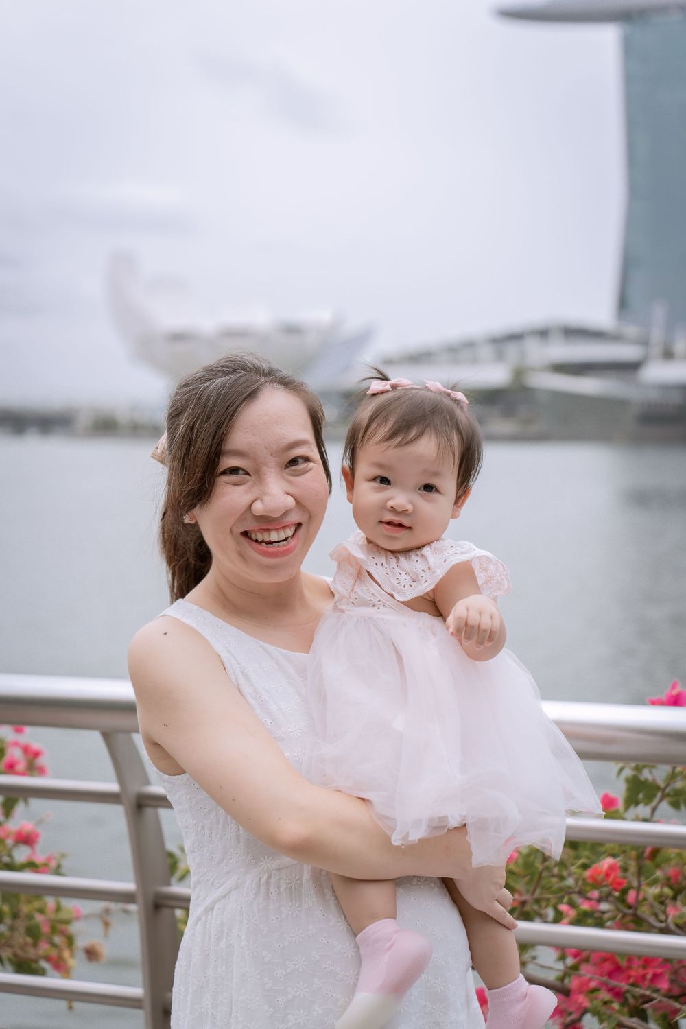 Parent and baby in matching white outfits pose by Singapore waterfront with iconic hotel in background.