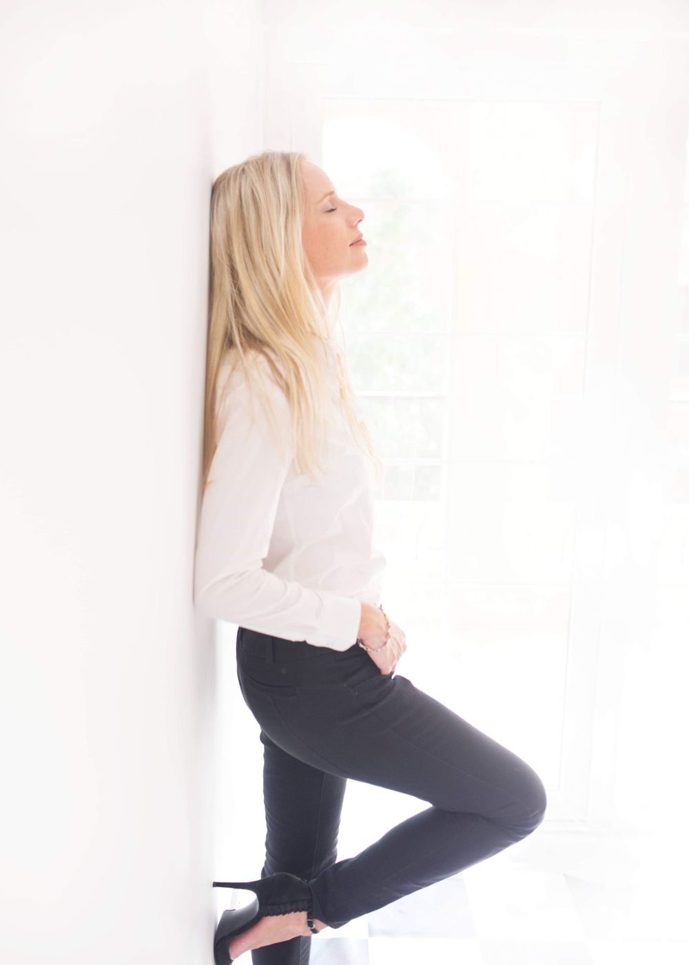 Woman in white long sleeve top and dark pants leaning against white wall in relaxed pose.