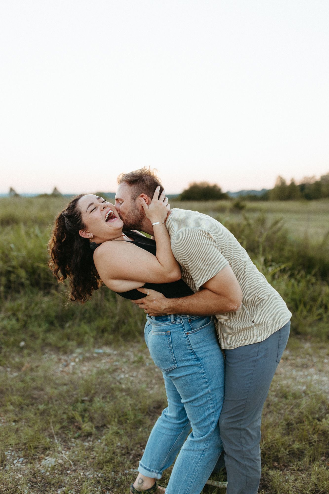 Playful couple embracing and laughing together in field during golden hour.