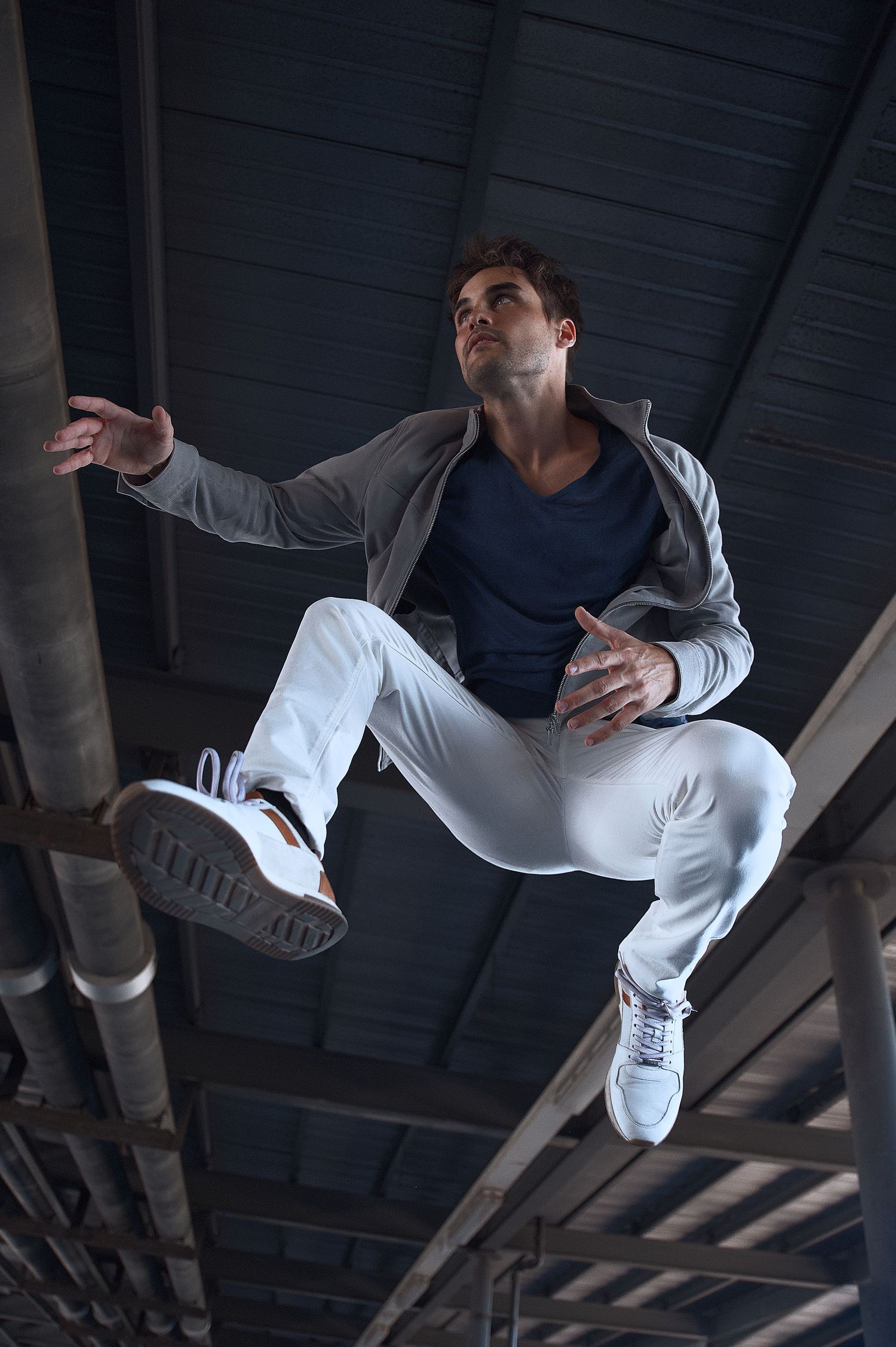 Modeling poses for male: An athletic model in white jeans and a grey jacket performs a dynamic jumping pose under an industrial ceiling.