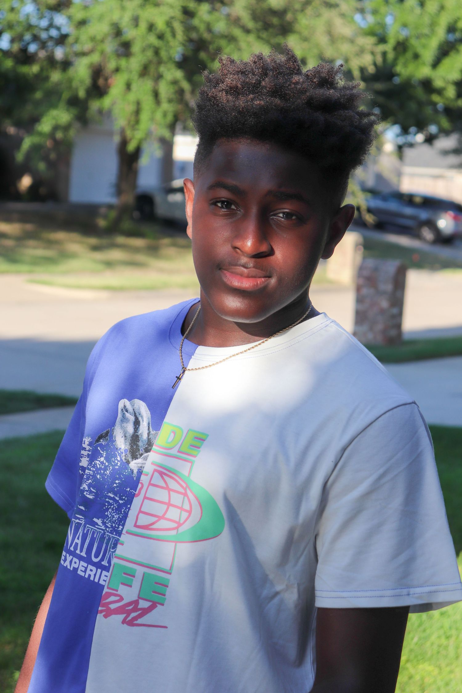 Young person in a white graphic t-shirt poses outdoors on a sunny day with trees in the background.