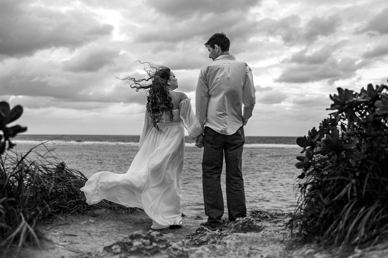 A couple stands together looking out at the ocean with dramatic cloudy skies.