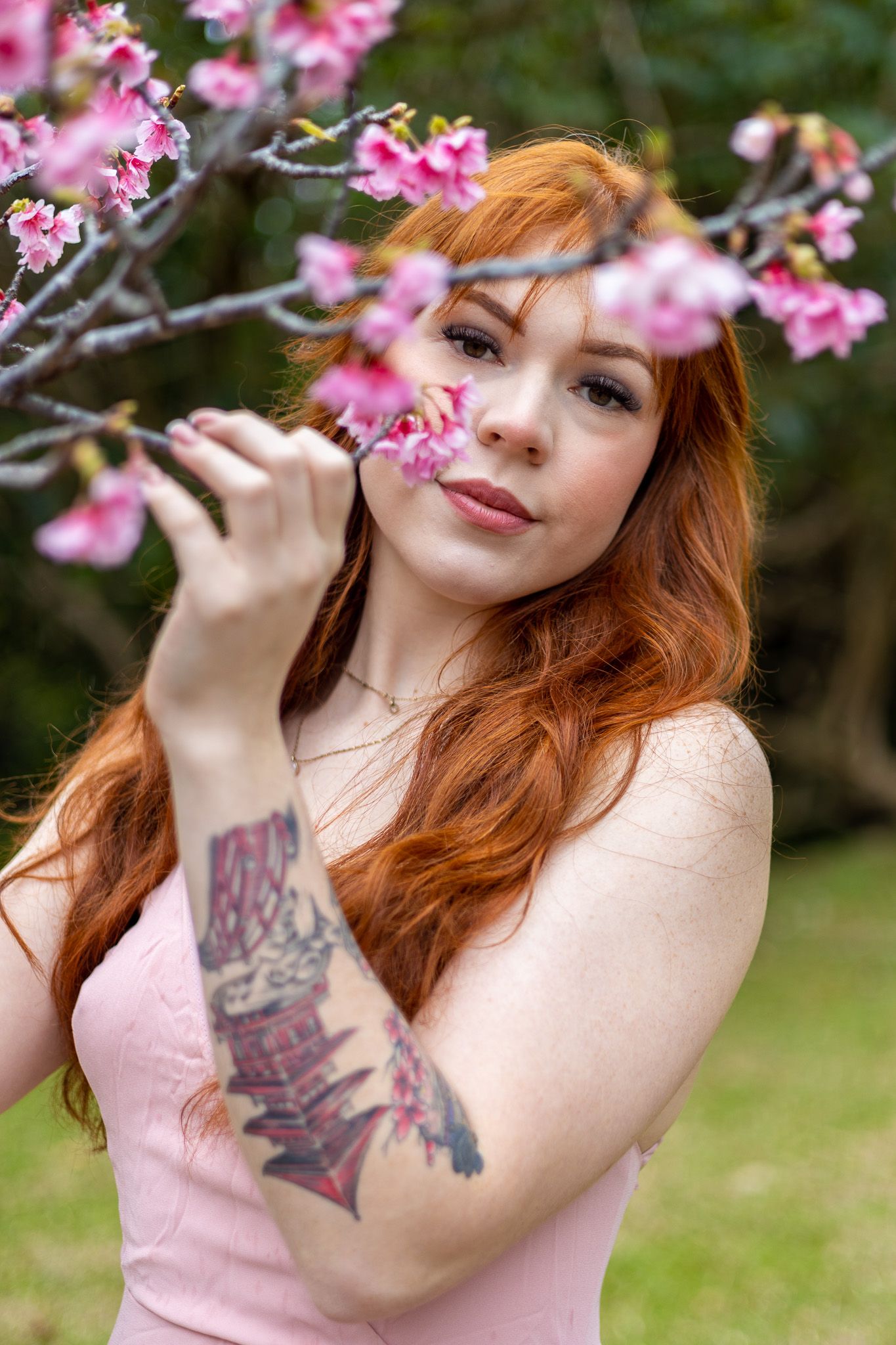 Person with tattoos and pink dress poses with cherry blossoms in spring garden.