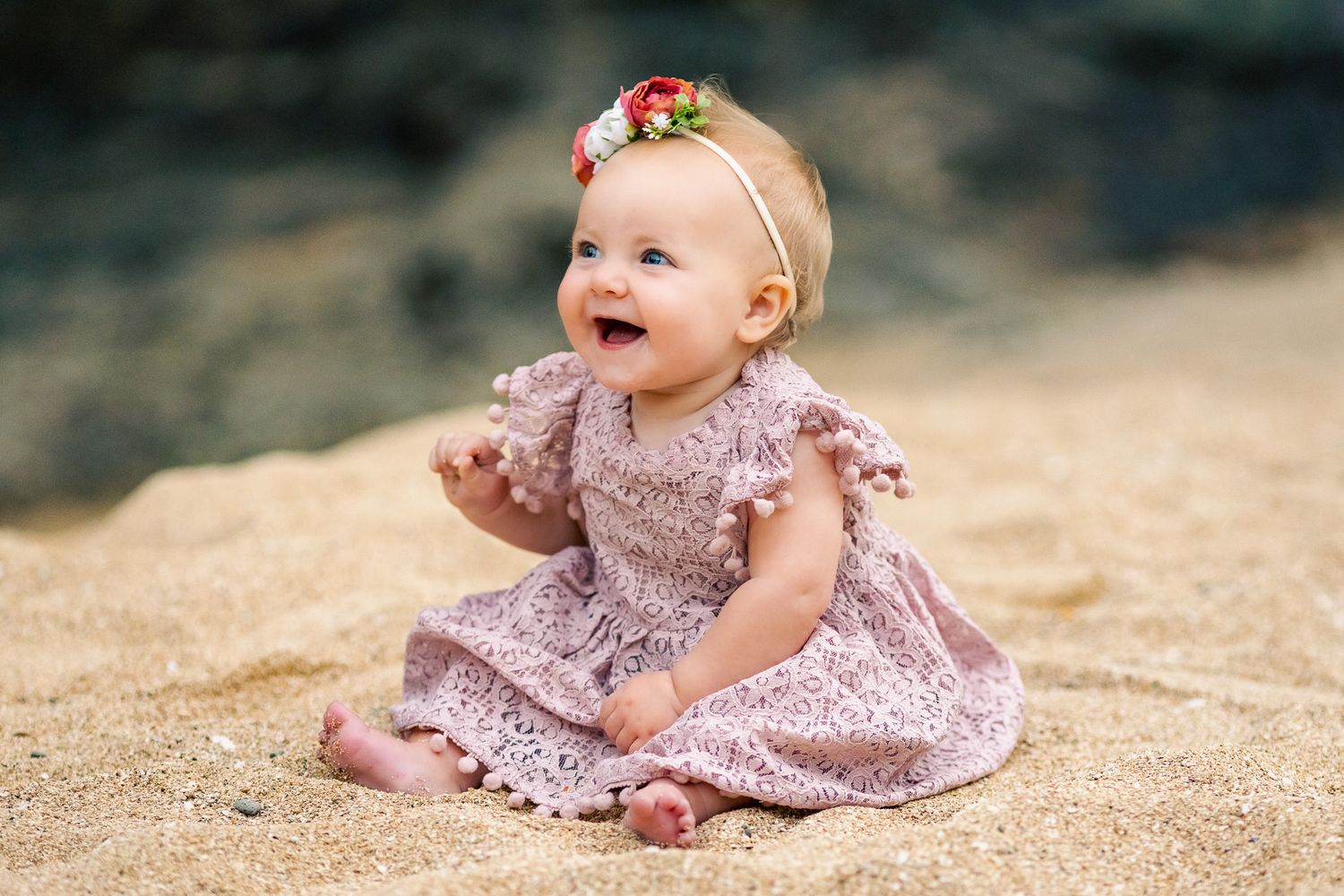 Baby in pink dress sitting and laughing on sandy beach.