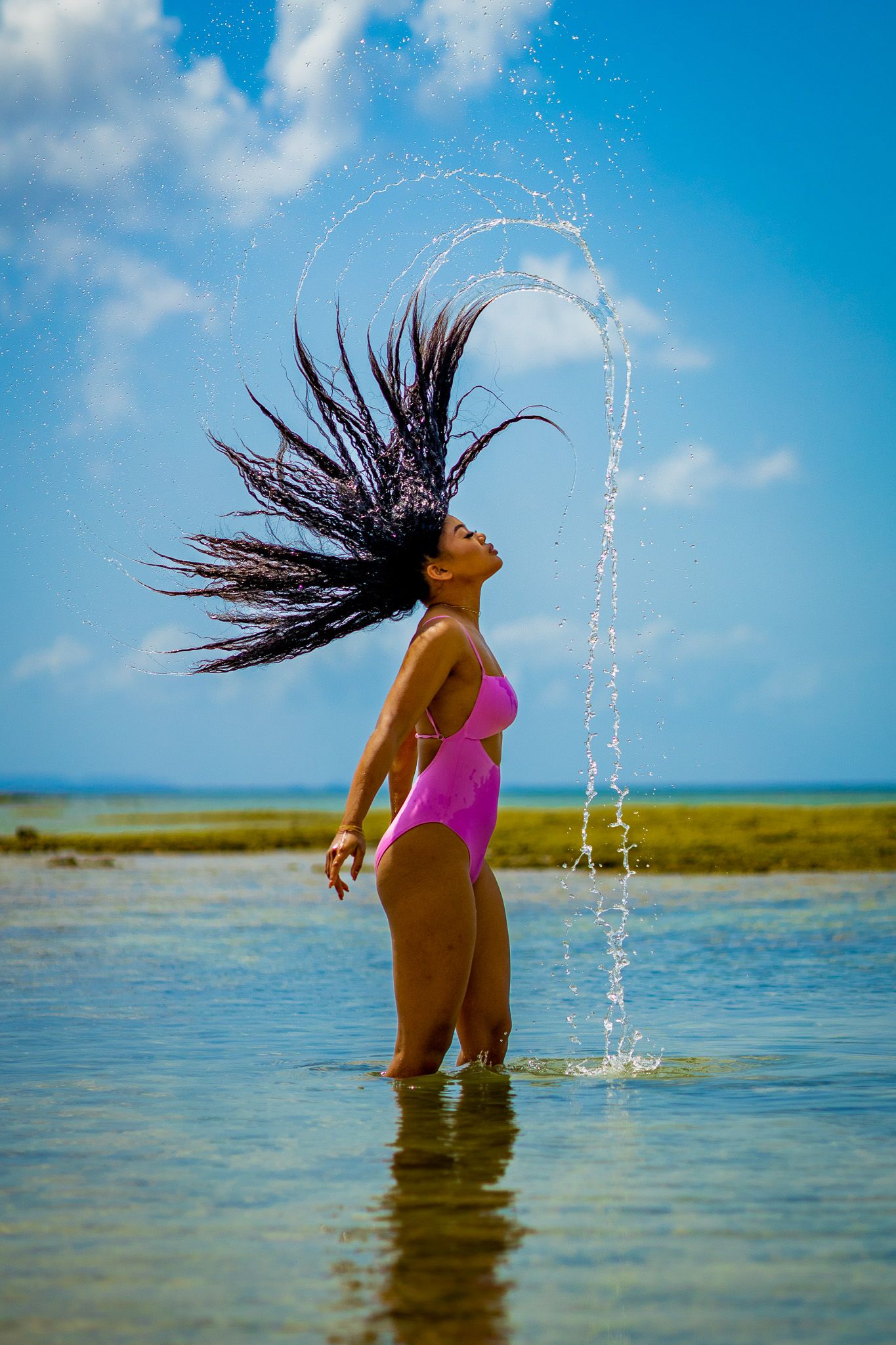 A person in a pink swimsuit flips their hair back in shallow water against a bright blue sky.