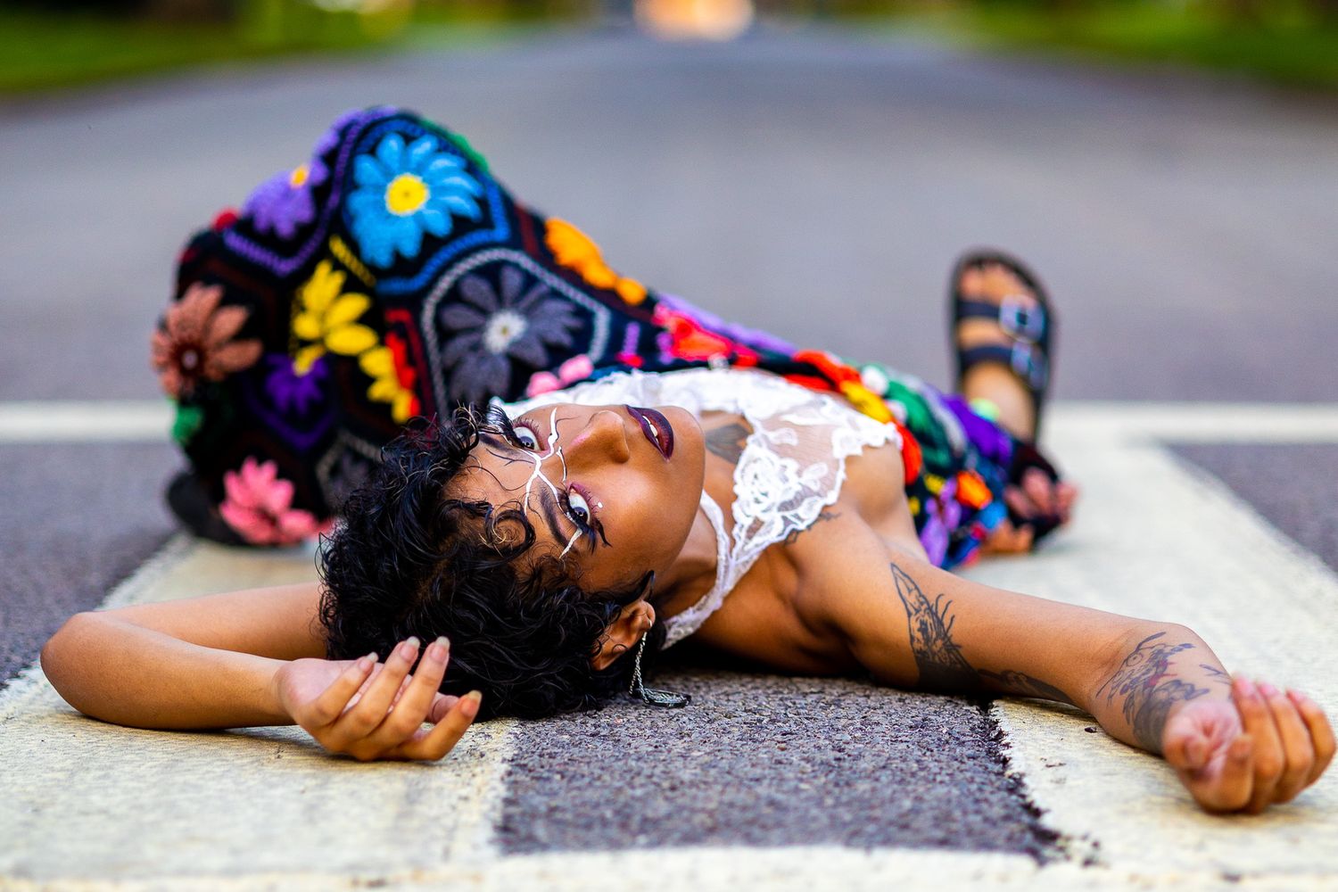 Person lying on pavement wearing colorful floral pants and white top in artistic pose.