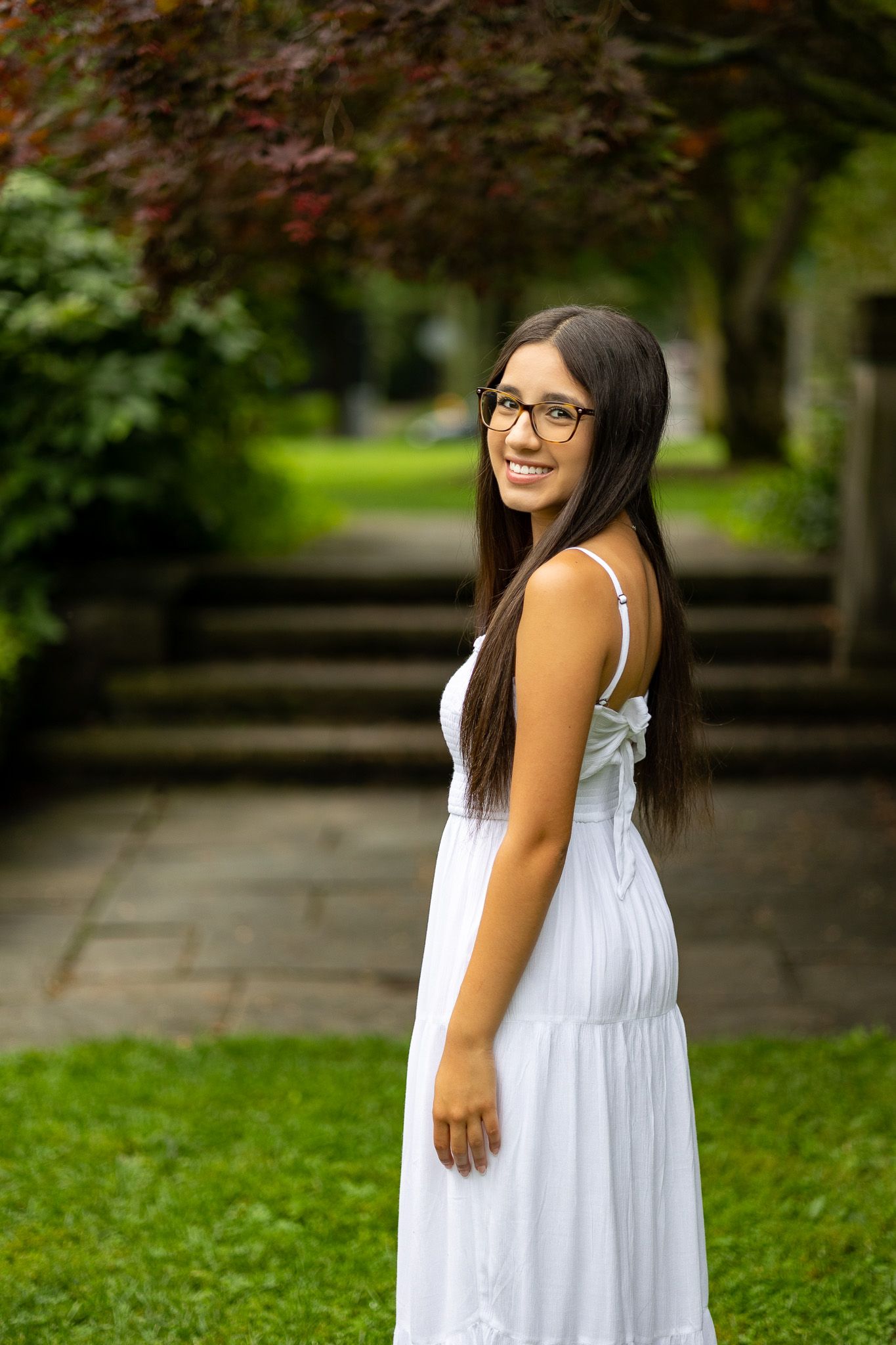 Young woman in white summer dress smiling in garden setting with trees in background.