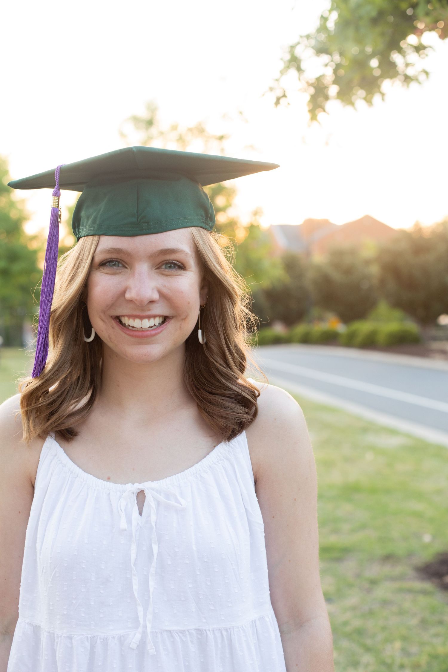 Graduate wearing a cap and white dress stands outdoors during golden hour.