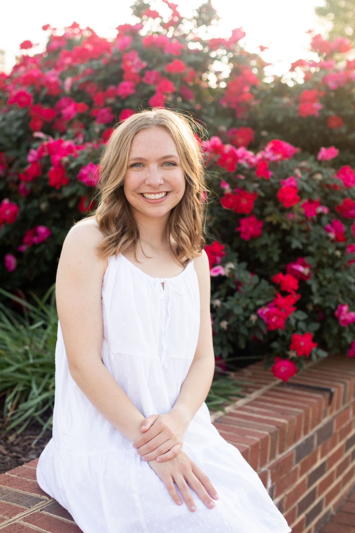 Graduate in a white dress poses in front of vibrant red roses blooming on a brick wall.