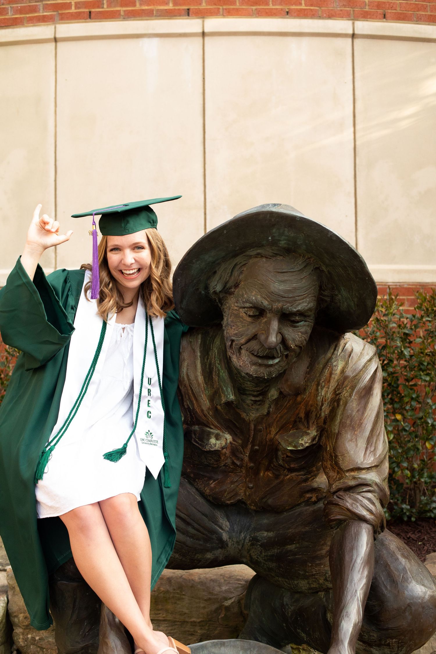 Graduate in green regalia poses playfully next to bronze statue on campus.