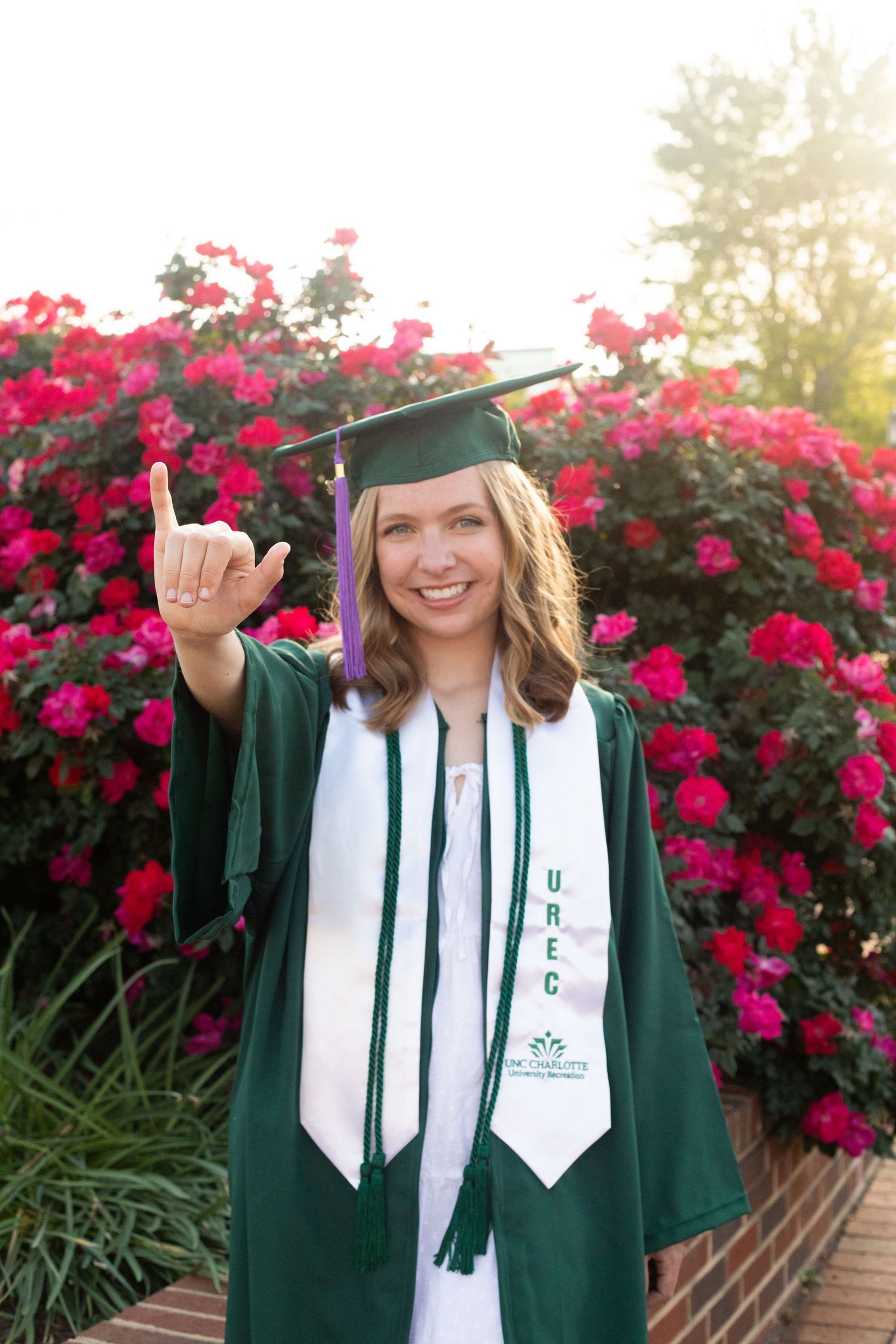 Graduate in green cap and gown makes shaka sign against backdrop of blooming red flowers.