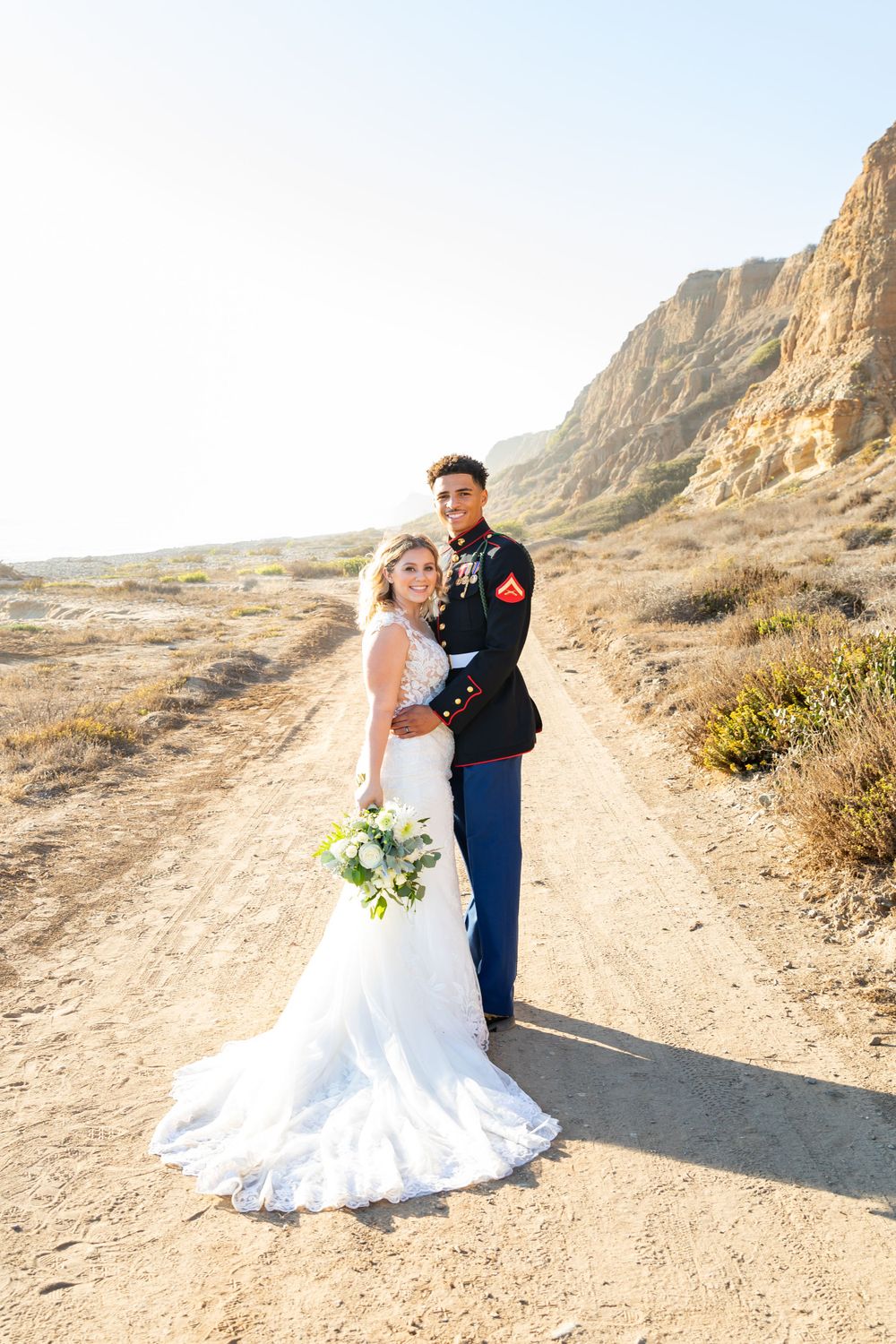 Newlyweds stand together on a desert path with stunning cliff formations behind them.