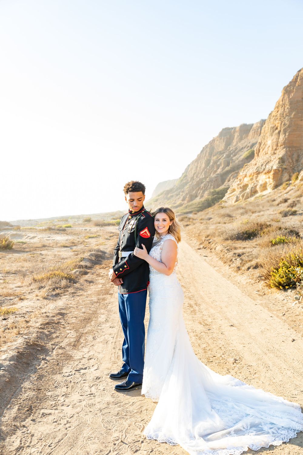 A military couple poses for wedding photos on a desert trail with towering cliffs.
