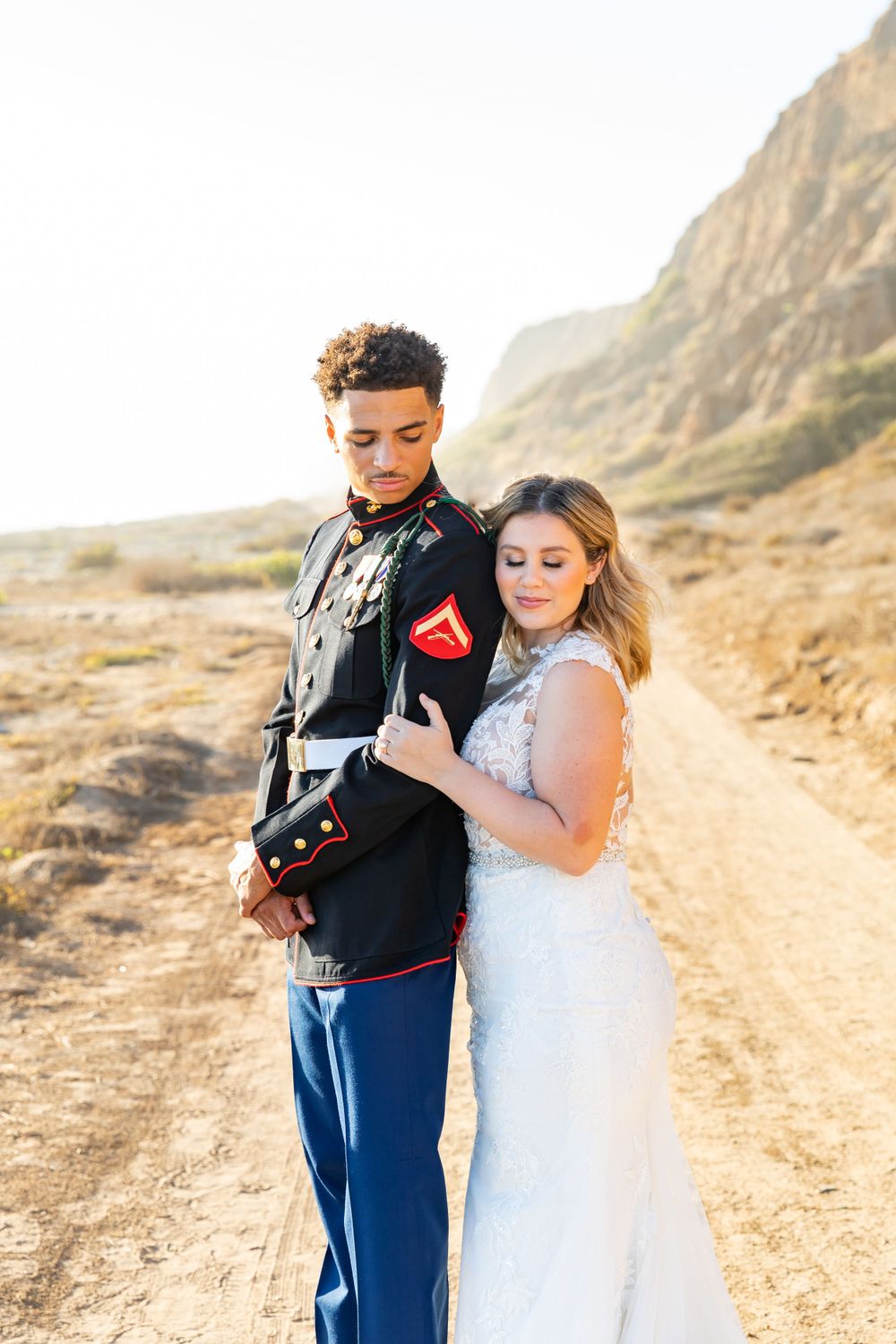 A Marine in dress blues and bride in lace gown pose together on a desert path.