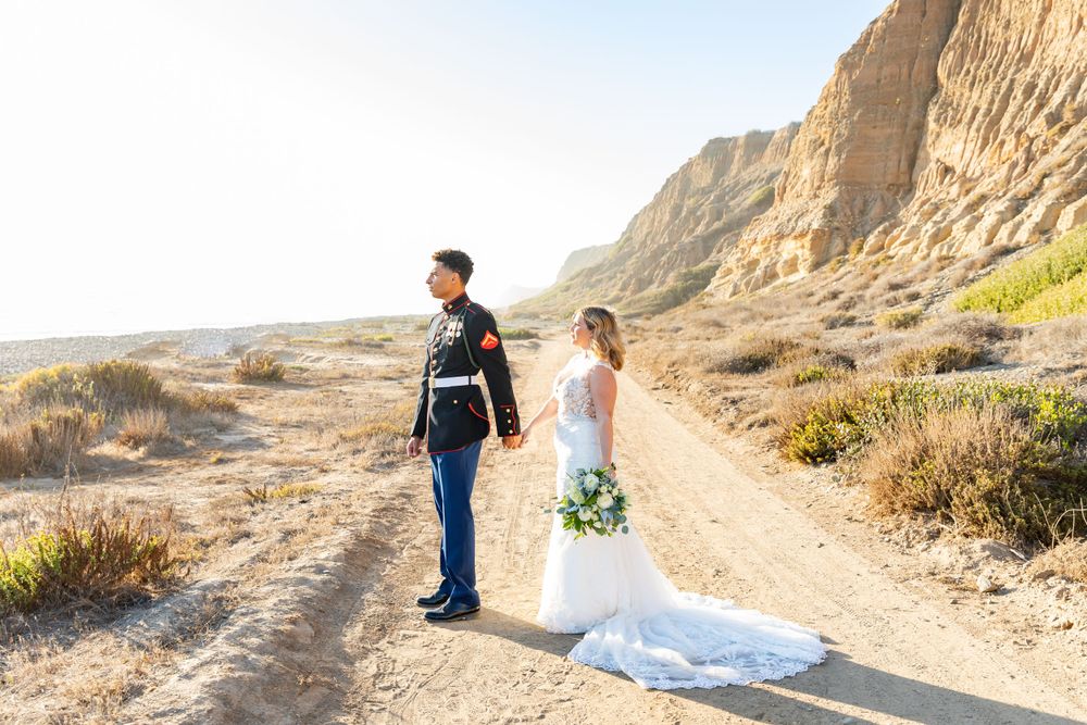 Newlyweds walking hand in hand on a desert path with dramatic cliff backdrop.