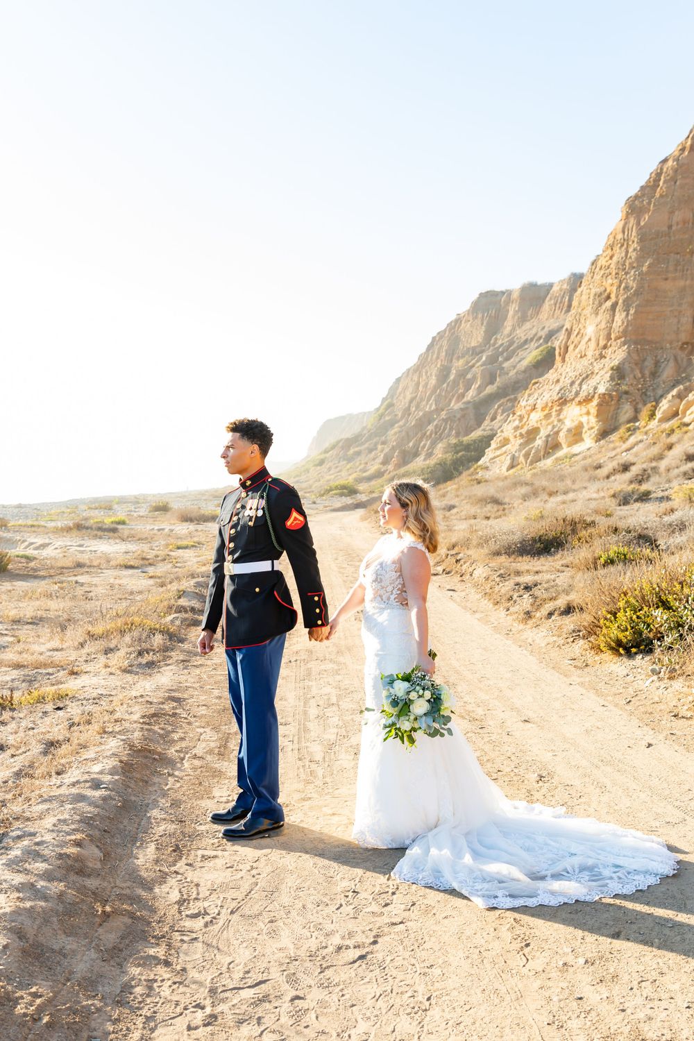 A military wedding couple stands on a desert trail with majestic cliffs in golden light.