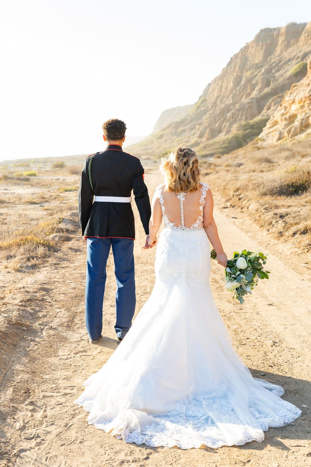 Military couple walking away showing beautiful bridal gown train against desert landscape.
