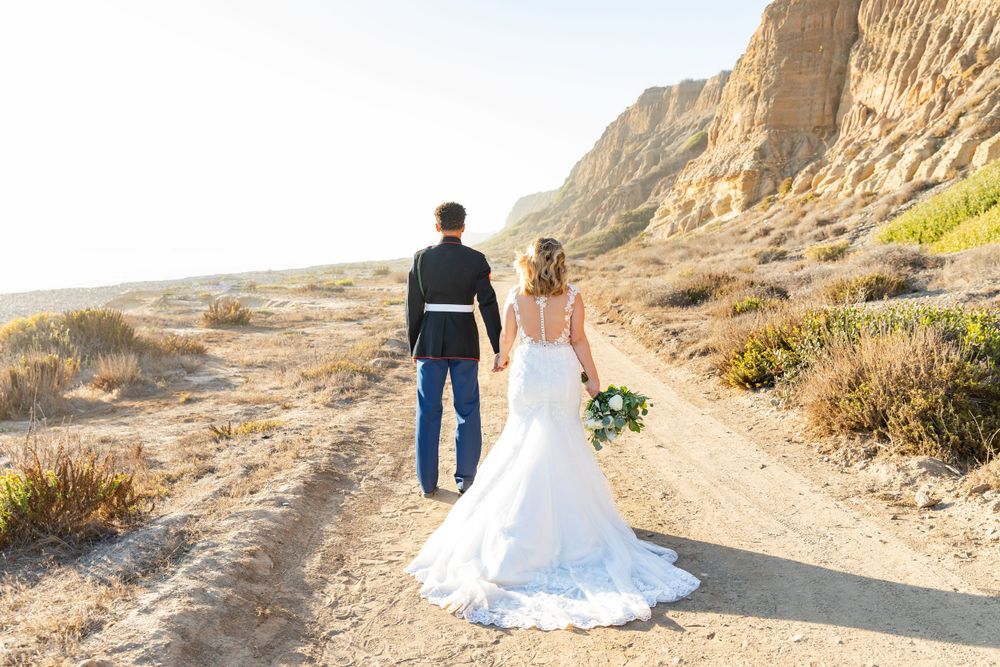 Bride in flowing white gown and serviceman walk along desert trail with cliffs in background.