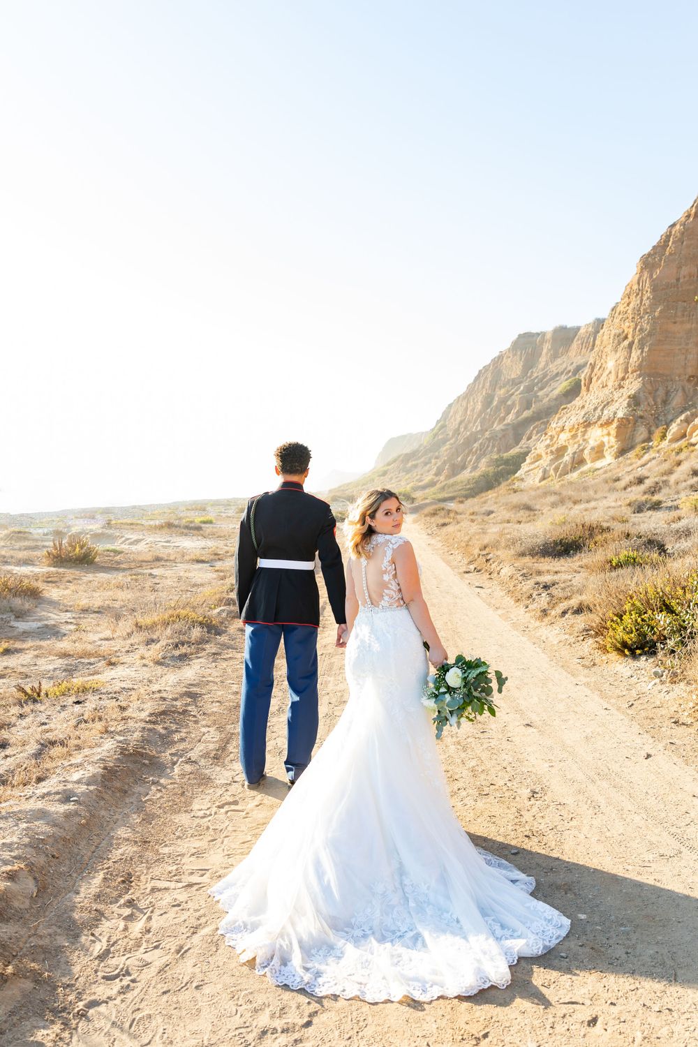 Wedding couple poses on dirt path with majestic desert cliffs creating a stunning backdrop.