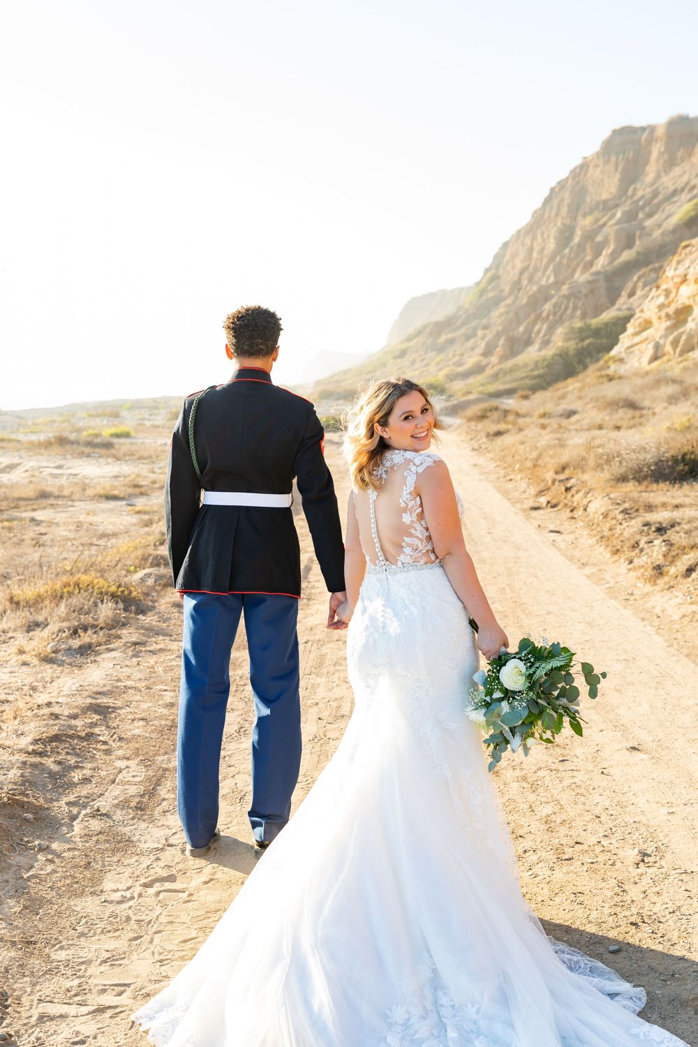 A military service member and bride walk along a scenic desert trail with dramatic cliffs in the background.