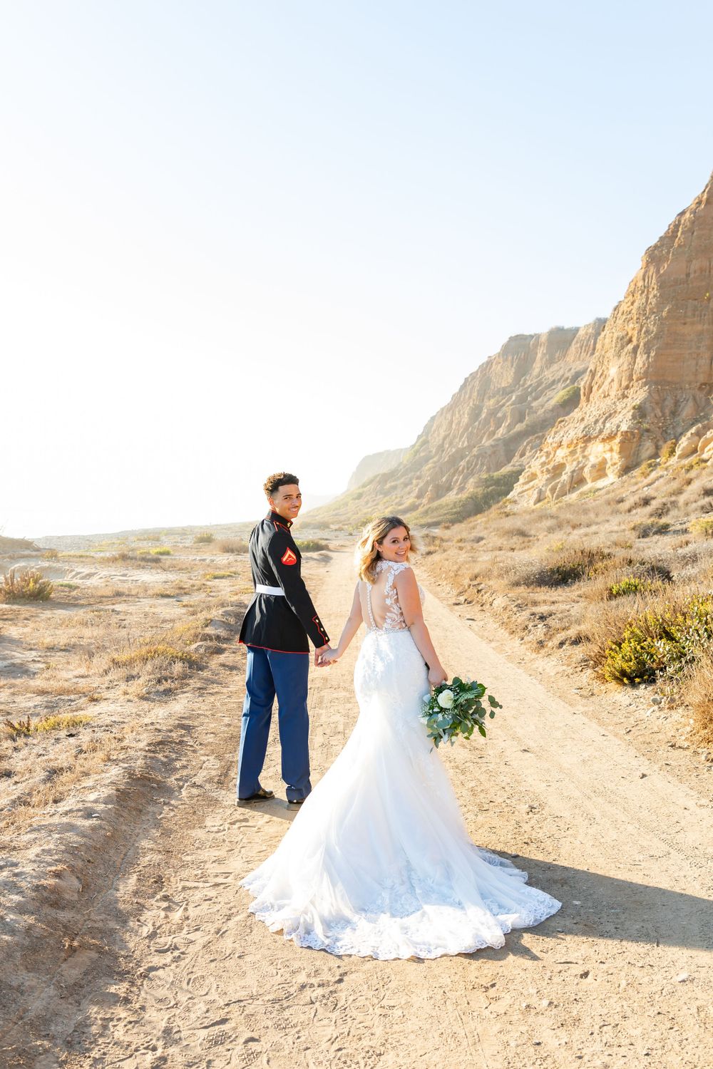 Newlyweds hold hands while walking on desert trail with rocky cliffs and golden sunlight.
