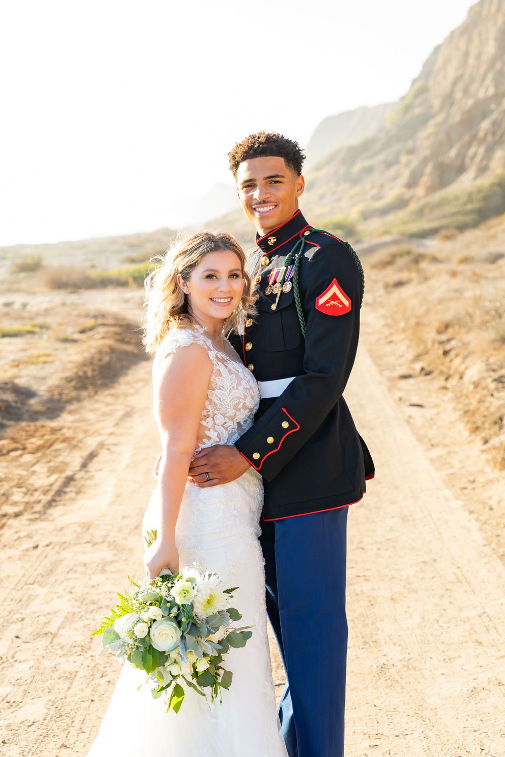A military wedding couple poses on a scenic dirt trail with dramatic cliffs in the background.
