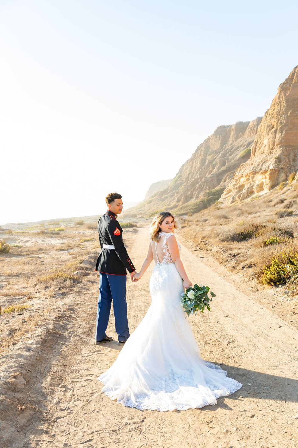 Military groom and bride in lace dress pose together on desert path during sunset.