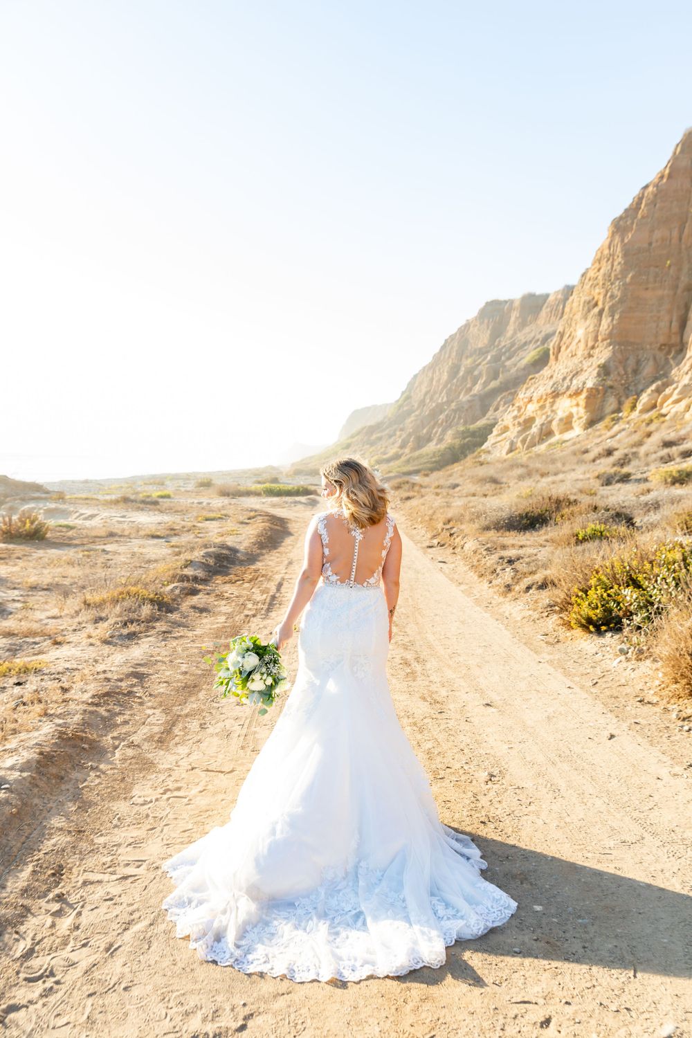Bride in flowing lace gown walks along desert path carrying fresh bouquet.