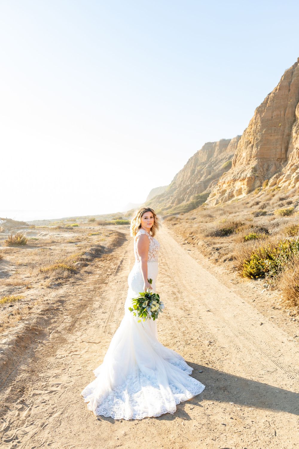Elegant bride in white gown stands on desert trail with mountains in background.
