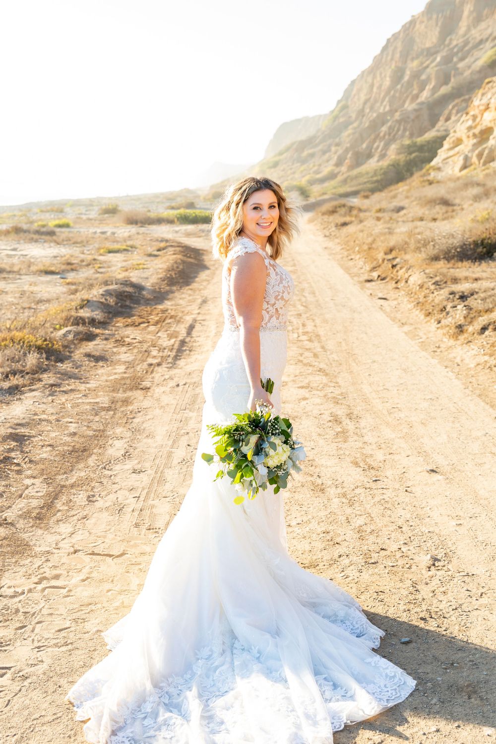 Bride poses in fitted lace wedding dress holding greenery bouquet on desert path.