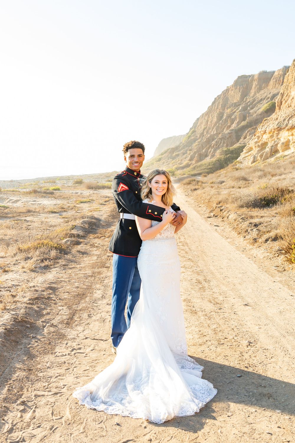 Military groom twirls bride in white dress on scenic desert path.