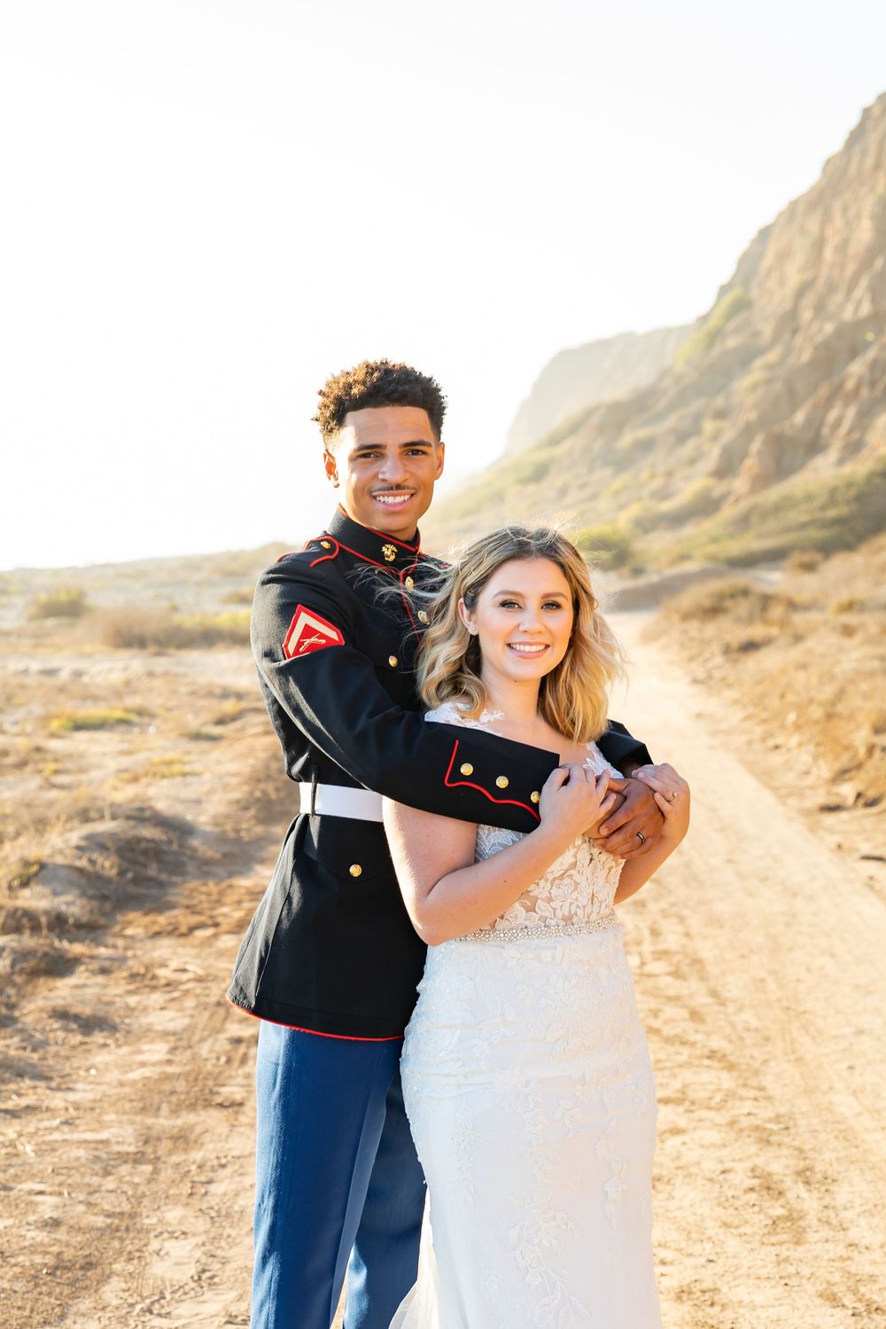 A military servicemember and bride embrace on a scenic desert trail with rocky cliffs in the background.