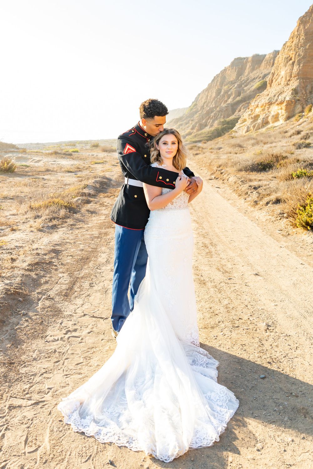 A newlywed couple in military dress uniform and lace wedding gown stand together on a sunlit desert path.
