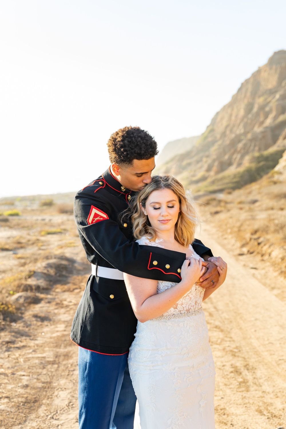 A romantic embrace between a military groom and bride on a desert mountain trail.