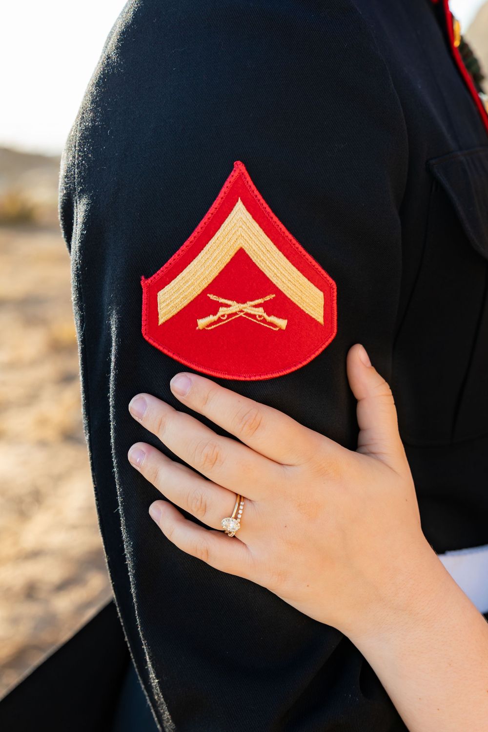 Close up of a Marine Corps Lance Corporal rank insignia patch on a dark uniform with a hand wearing an engagement ring.