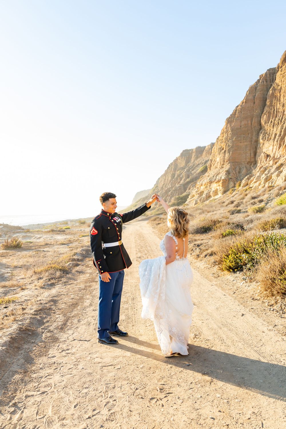 A military service member and bride dance on a desert trail with dramatic cliffs in the background.