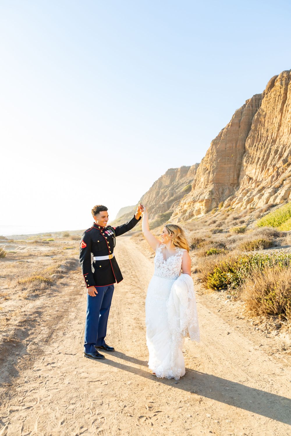 A couple in military dress blues and wedding gown twirl on a scenic desert path with towering cliffs.