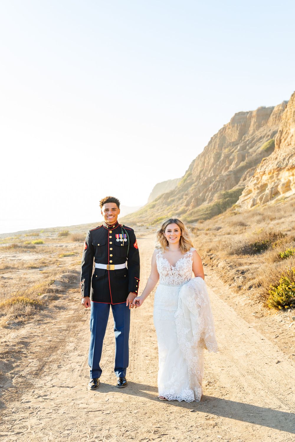 A military wedding couple walks hand in hand along a desert trail with majestic cliffs in the background.